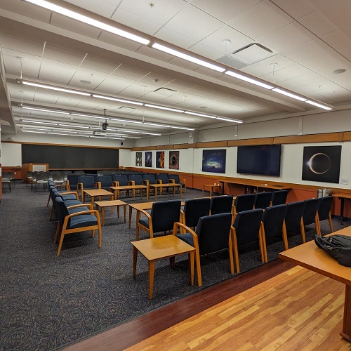 An interior room with numerous chairs arranged to face a central area; behind them, chairs facing a blackboard with a podium.