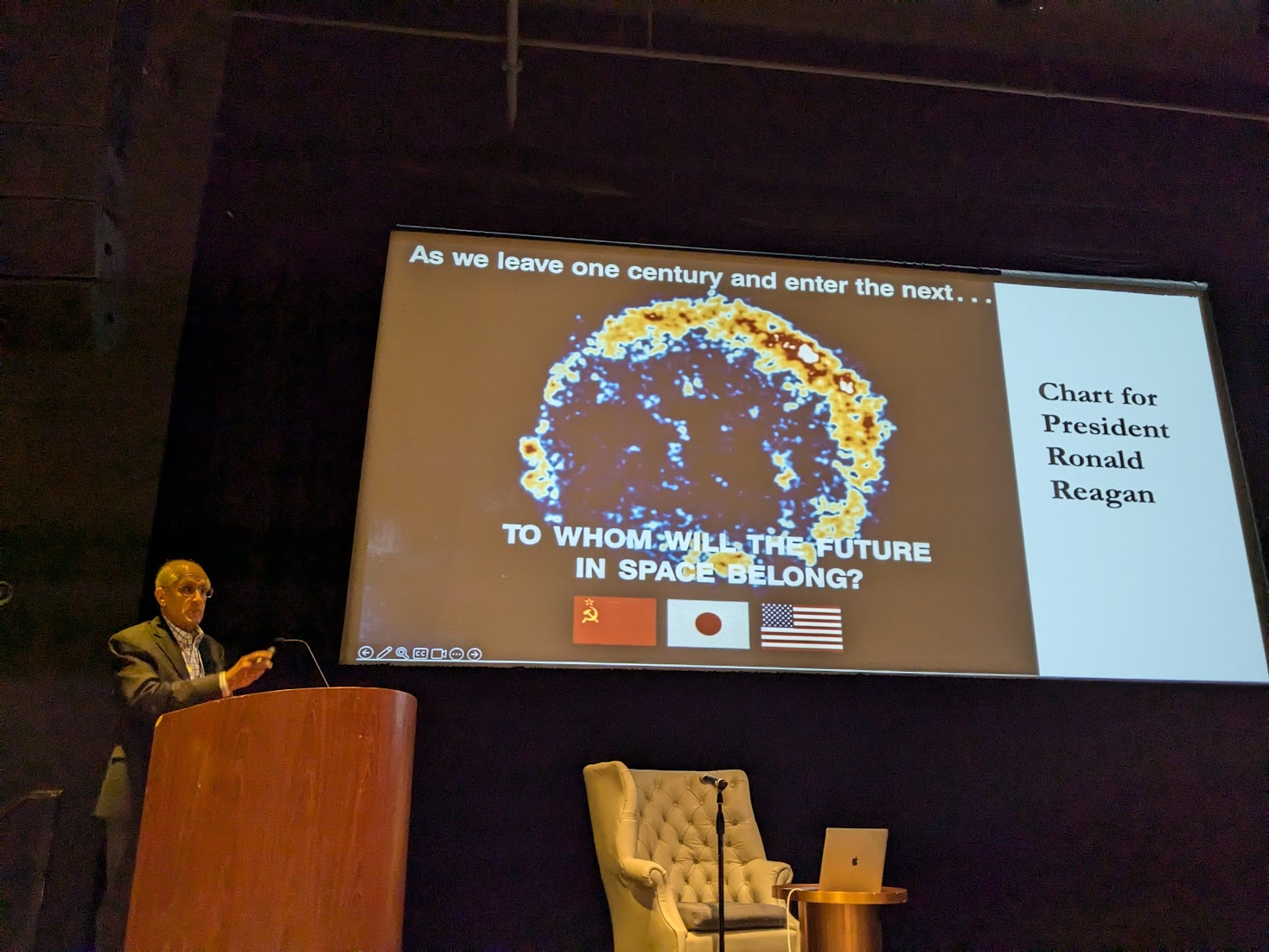 Harvey Tananbaum stands at a lectern in front of a projected slide. This slide shows a crude image of a supernova remnant with the words 'As we leave one century and enter the next⦠to whom will the future in space belong.' Underneath are the Soviet, Japanese, and American flags. At the side of the slide is the caption 'Chart for President Ronald Reagan.'