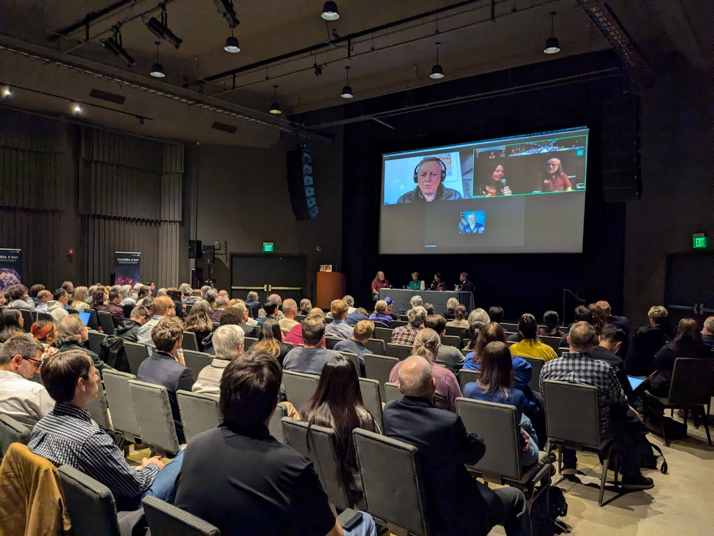 Chandra X-ray Center director Pat Slane sits at a table at the front of a spacious theater alongside three astronauts: Eileen Collins, Catherine Coleman, and Jeffrey Ashby. Above them is a displayed Zoom window, showing Michel Tognini. The audience is visible, with overflow seats occupied.