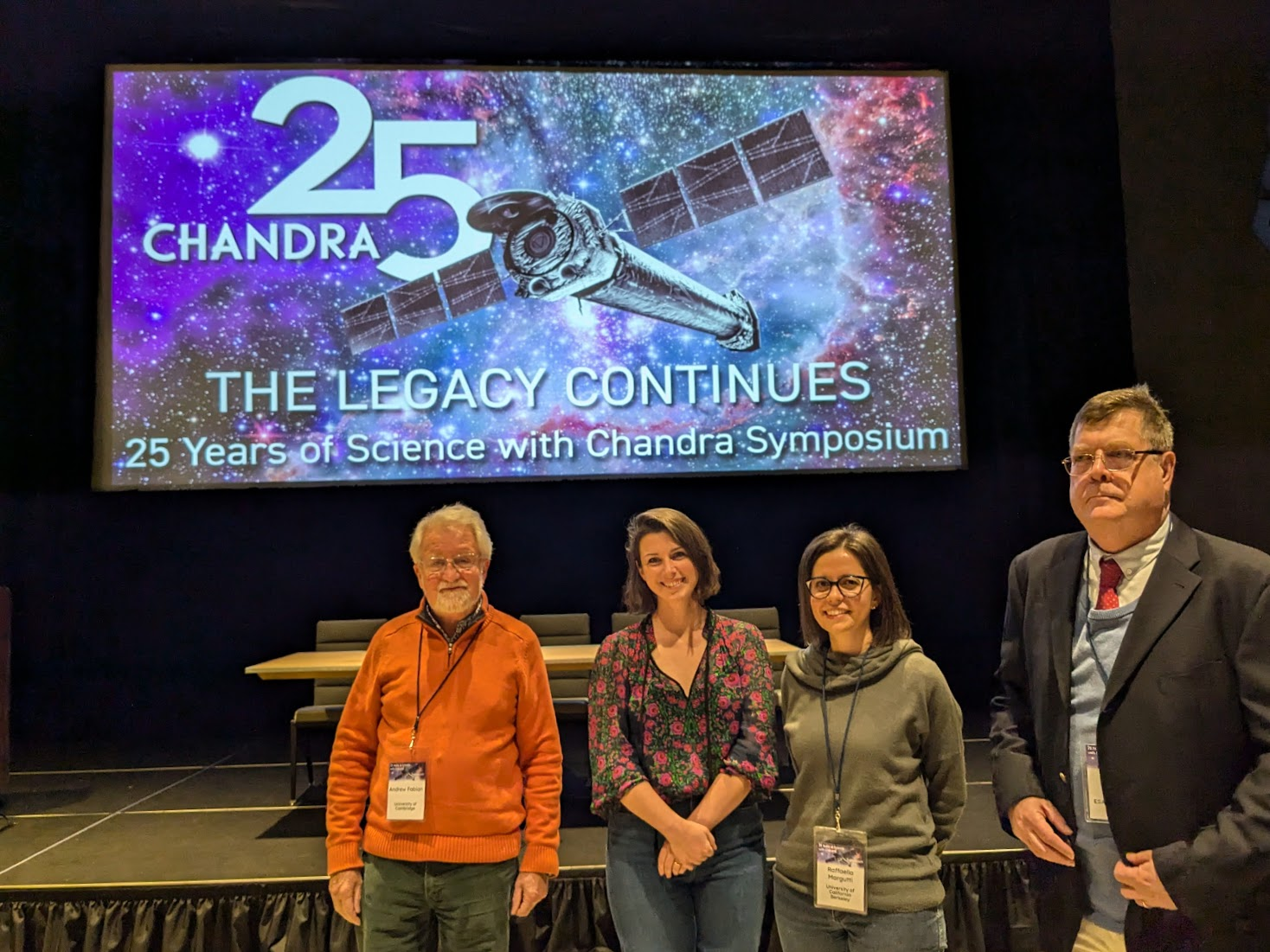 Four people stand in front of a raised stage with a long table set upon it. Behind them is a projected screen showing the symposium logo for the Chandra 25 symposium.